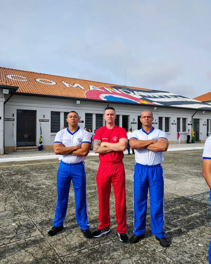 Kilder, à direita, com dois colegas de uniforme de educação física da PM, de braços cruzados em frente ao prédio do comando