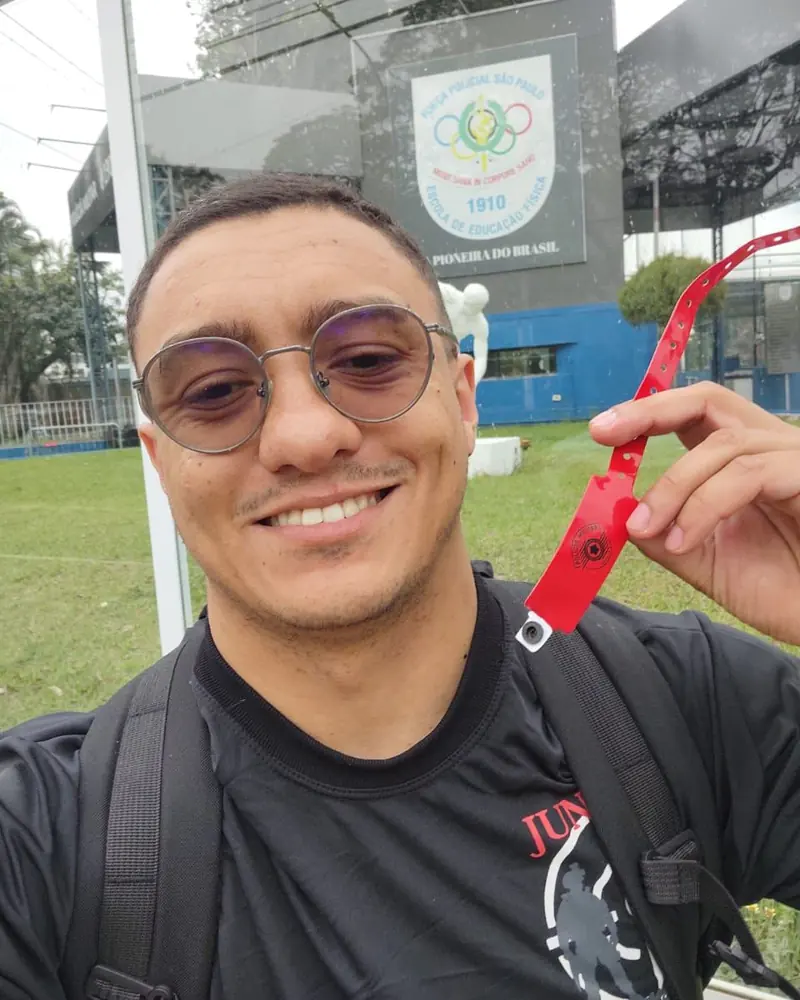 Antes: o aluno sorrindo, de camiseta preta do Juntos, mostrando a pulseira vermelha da Polícia Militar em frente à Escola de Educação Física