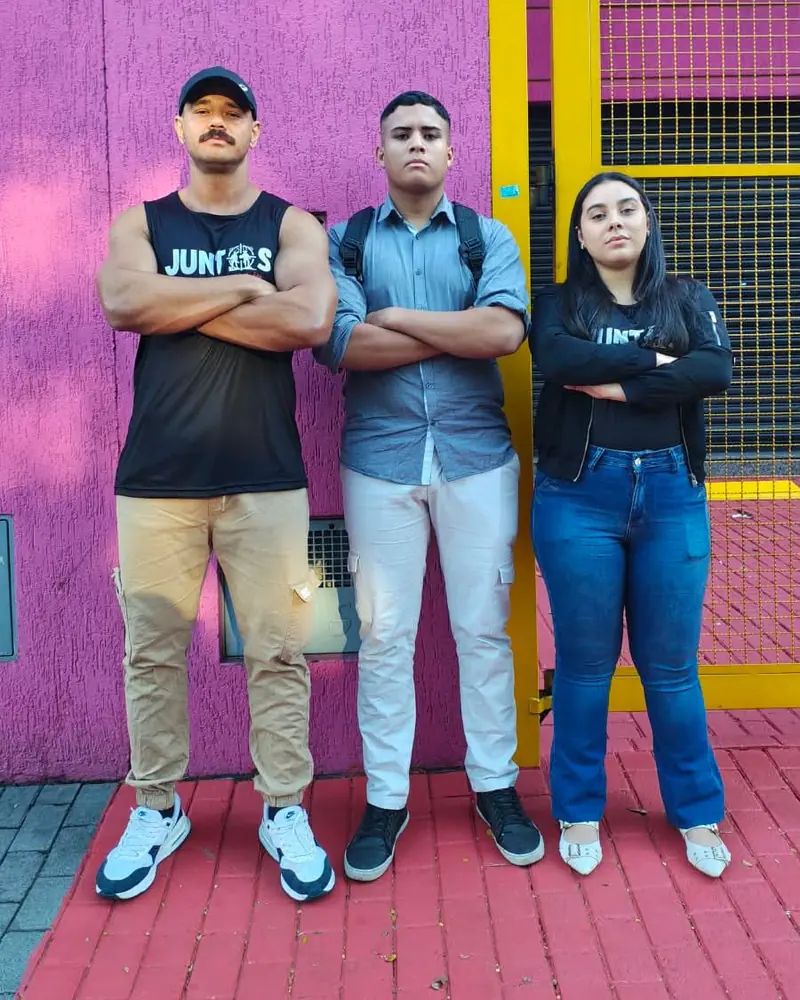 Três alunos do Juntos de braços cruzados em frente a um muro rosa, dois com a camiseta preta do Juntos